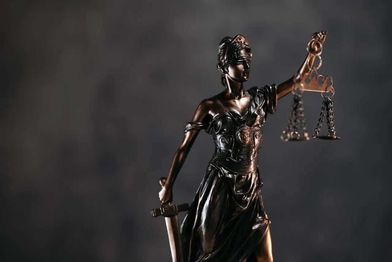 “Bronze statue of Lady Justice holding a sword in one hand and balanced scales in the other, wearing a blindfold to symbolise impartiality, against a dark blurred background.”