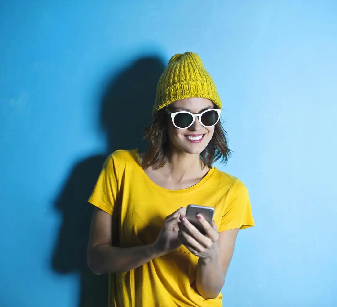 “Woman in yellow clothing and dark glasses navigating a mobile phone against a blue background”